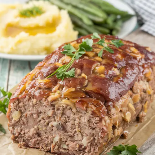 A photo of a golden-brown loaf of meatloaf with a caramelized brown sugar glaze on a rustic wooden cutting board. The meatloaf has a moist and tender interior flecked with onions and herbs. Fresh parsley is scattered over the top for a pop of color. On the side, there is a plate of buttery mashed potatoes and roasted green beans. The whole setup radiates the warmth of a classic family dinner. The title text overlay says "Classic Diner Style Meatloaf with Brown Sugar Glaze" in a soft feminine country font.