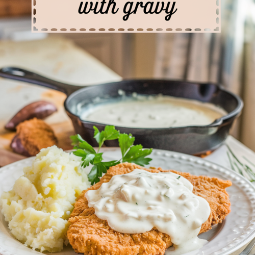 A warmly lit rustic kitchen with a country-style dining setting. The main focus is a classic country dinner scene of a golden-brown country fried steak topped with rich, creamy white sausage gravy. The plate is accompanied by fluffy mashed potatoes on the side, a sprig of fresh parsley for a touch of green, and a skillet filled with extra gravy placed nearby. The background contains a few kitchen items and a window with a curtain. The image has a title text overlay "Country Fried Steak with Gravy" in a soft feminine country font.