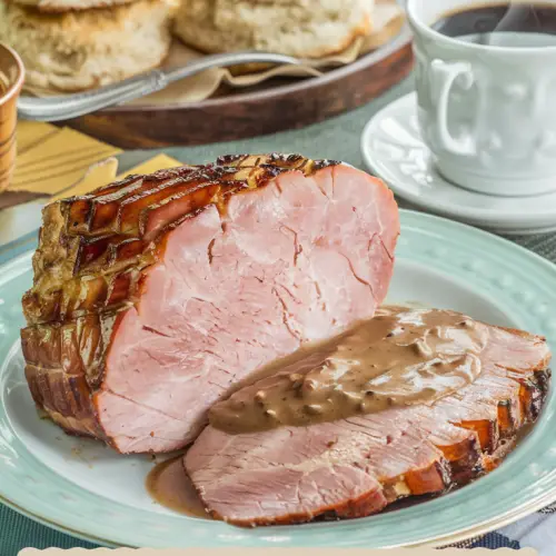 A photo of a plate with a thick-cut slice of country ham. The ham is seared to golden-brown edges and glistening with its savory juices. Next to the ham is a pool of rich, coffee-infused red-eye gravy. The background has a stack of fluffy ham biscuits on a wooden tray and a steaming cup of black coffee. The table is set with warm tones and a touch of Southern flair. Title text overlay "Country Ham and Red-Eye Gravy" is done in a soft feminine country font.