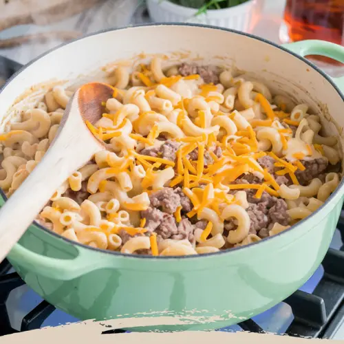 A cozy kitchen scene with a steaming pot of cheesy hamburger macaroni on a stove. The pot's creamy sauce is bubbling gently, with bright orange cheddar cheese melted over tender elbow macaroni and juicy ground beef scattered throughout. A wooden spoon rests in the pot, ready to serve, and a bowl of fresh parsley adds a pop of green to the warm, inviting scene. In the background, a loaf of crusty bread and a pitcher of iced tea complete the image of a cozy family dinner. The title text "One Pot Easy Cheesy Hamburger Macaroni" is overlaid in a soft feminine country font.